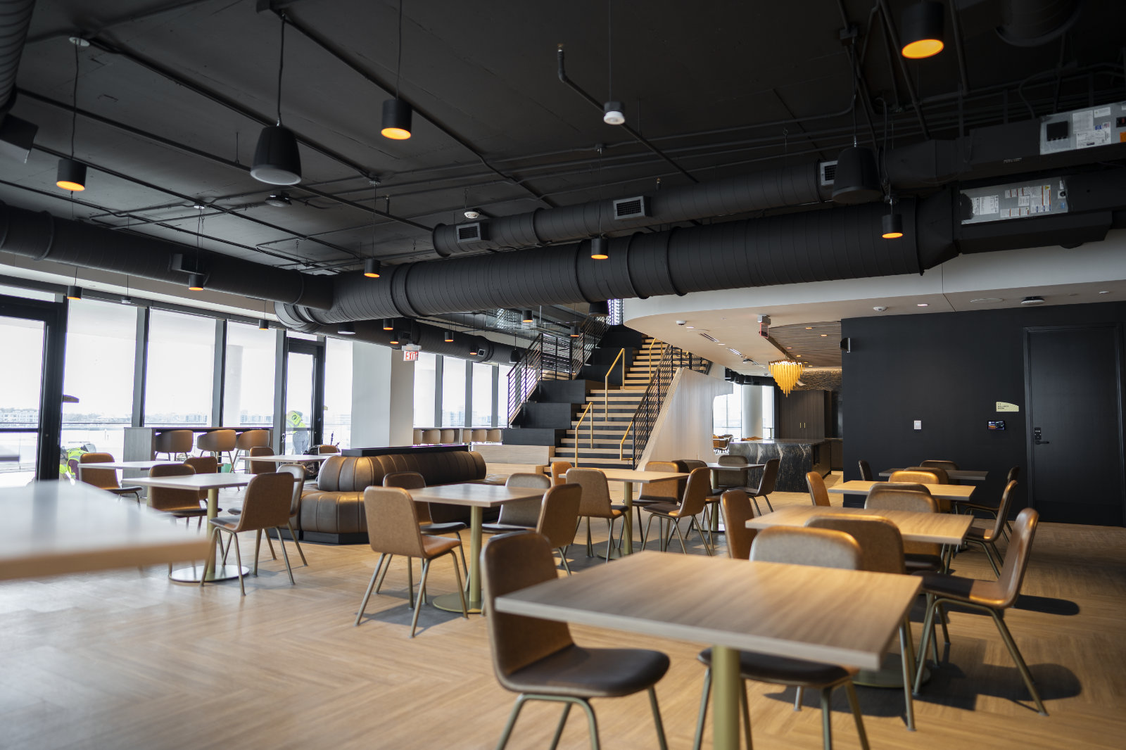 Softly lit lobby with square tables, chairs and hightop tables in light wood colors with black ceiling and floor to ceiling windows on left.