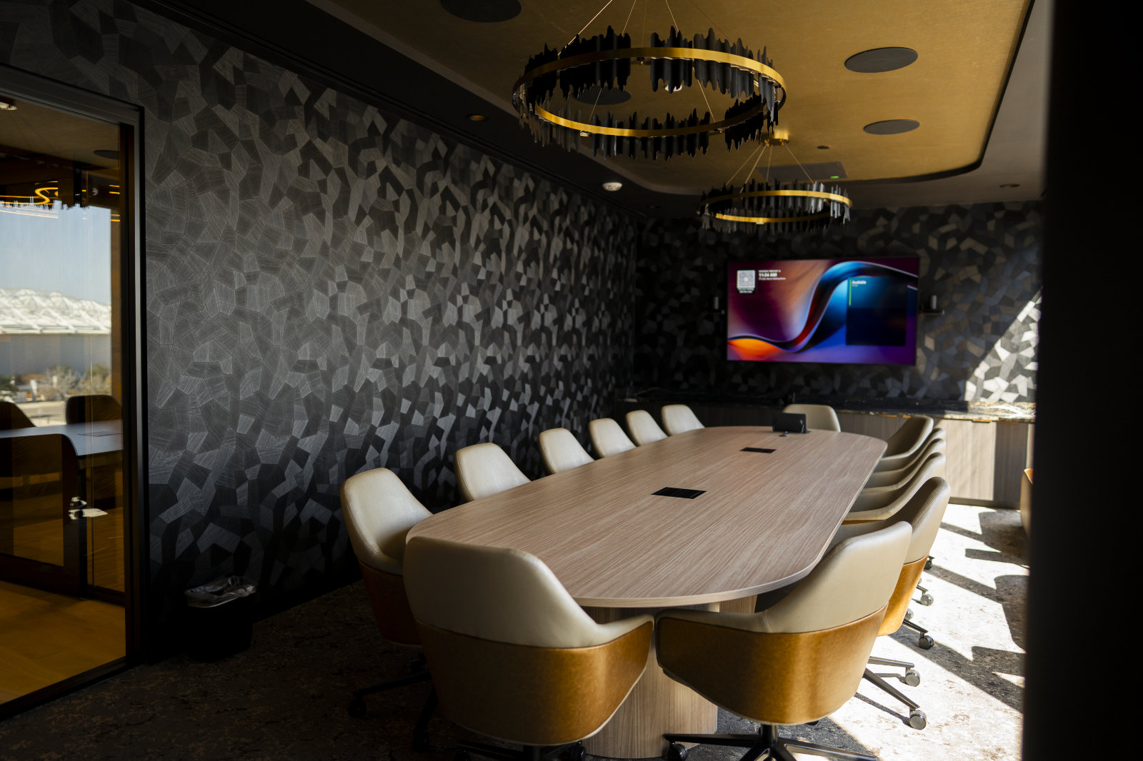 Conference room features long oval table and two circular chandeliers surrounded by dark gray and black textured wall paper with a colorful TV display at the end.