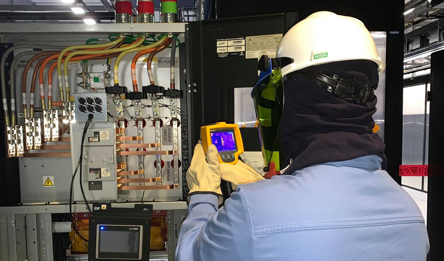 Technician wearing protective gear uses a handheld thermal camera to inspect electrical switchgear and power cables inside an industrial facility.